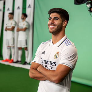 Marco Asensio, soccer player, poses confidently with a smile. White jersey with Adidas and airline logos. Green field, blurred background, other player's legs visible.