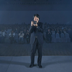 Masami Kawasaki, Canon executive, presenting Canon EOS 7D Mark II at press conference. Smartly dressed in black suit, holding camera. Crowd in background.