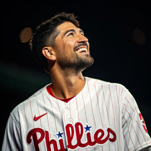 Nick Castellanos, Hispanic baseball player, beams joyfully in a high-definition photo shoot. He's clad in a white and red Philadelphia Phillies jersey adorned with the Nike swoosh. Dark backdrop highlights his vibrant expression and attire.
