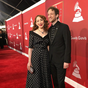 Regina Spektor and Jack Dishel at the Grammy Awards ceremony. Wearing a black polka dot dress and a formal black suit. Red carpet setting with vibrant red backdrop and music-related logos.