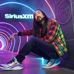 Steve Aoki, Asian musician with long hair and beard, posing in a neon-lit venue. He's wearing a vibrant tartan shirt, dark jeans, and futuristic sneakers. The background is a swirling vortex of colors with the SiriusXM logo prominently displayed.