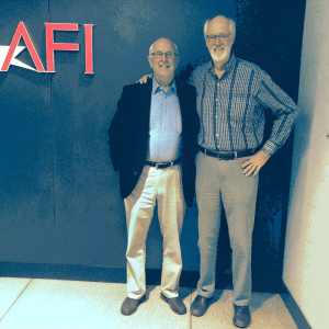 James Cromwell and John Cromwell at an awards show, James in a dark blazer, button-up shirt, and belt. John in a plaid shirt, belt, casual stance. Wall features bold red AFI logo. Even lighting, friendly atmosphere.