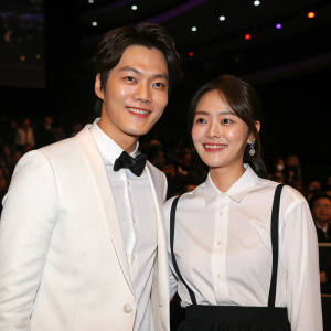 Joo Won and Choi Gang-Hee, smiling at the KBS Drama Awards. White shirt and black skirt attire. Dimly lit room with audience in background.
