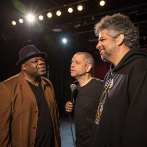 Patrice O'Neal, Jim Norton, and Greg Giraldo backstage at a comedy show. Patrice in a brown jacket, Jim in a black shirt, Greg in a graphic tee. Bright stage lights in background.