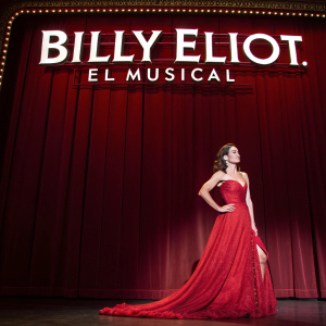 Natalia Millán in a stunning red gown, posing elegantly on the stage of a Broadway theater. The backdrop showcases the 'Billy Elliot. El Musical' title in vibrant white letters against a deep red curtain.
