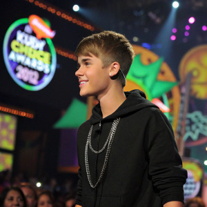 Justin Bieber, wearing a black hoodie and silver chain, at the Nickelodeon Kids' Choice Awards. Crowded stage with vibrant logos and colors in the background.