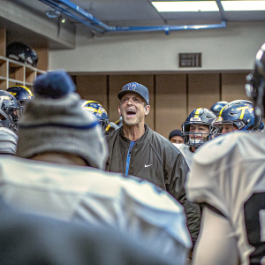 Jim Harbaugh in the center of a crowded locker room. Players and coaches in football gear surround him.