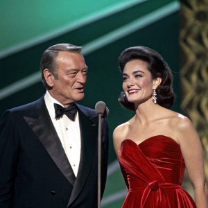 John Wayne and Pilar Palette Wayne at a glamorous award show. John in a sleek tuxedo, Pilar in a stunning red velvet gown. Both look elegant. The stage backdrop is a deep emerald green.