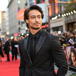 Tiger Shroff at movie premiere. Wearing black suit, tie, no beard. Crowded street backdrop.