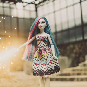 Lily Shimmer, a magical character from Monster High, with long, vibrant blue and pink hair, standing in a sparkling studio. Pale skin, wearing a colorful dress with geometric patterns.