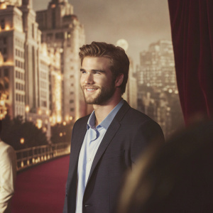 Liam Hemsworth at a gala event. Wearing a navy blazer and light blue shirt. No beard, clean cut hairstyle. Surrounded by bustling cityscape in the background