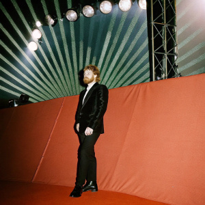 Ed Sheeran, Caucasian male with beard, at Grammy Awards. Wearing black suit and white shirt. Bright stage lights and backdrop of fan screens.