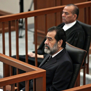 Former Iraqi President Saddam Hussein in a high-profile trial. Dark suit, beard. A man in similar attire sits beside him. Wooden courtroom decor.