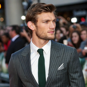 Alex Pettyfer, dressed in a sharp grey pinstriped suit, a crisp white shirt, and a dark green tie, captured during a candid moment at a bustling public event with a lively crowd in the background.