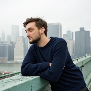 Richard Harmon, a man with a beard and short hair, in a casual photo shoot. Dark blue sweater. City skyline backdrop