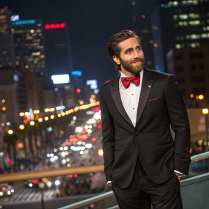 Jake Gyllenhaal, wearing a black tuxedo with a red bow tie, at an awards show. He stands confidently in front of a bustling cityscape.