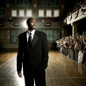 Kobe Bryant, wearing a black suit and tie, at a basketball hall of fame ceremony. A large, cheering crowd in the background.