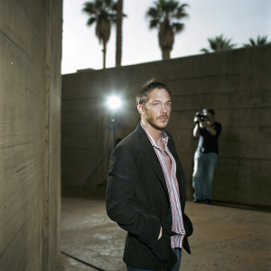 Tom Hardy, Caucasian actor with stubble and short hair, candidly captured outdoors. Dark blazer and striped shirt accentuate his rugged charm. Background blurred with photographer and palm tree. Golden hour lighting highlights his features.