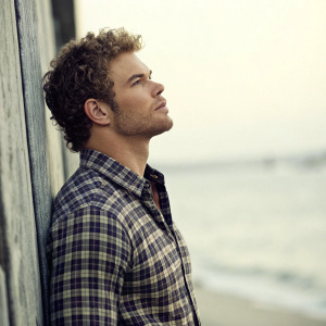 Kellan Lutz, a Caucasian actor with curly hair, stands against a blurred backdrop. Wearing a plaid shirt, he poses for a formal portrait emphasizing his upper body and facial features. The serene mood is accentuated by soft, diffused lighting.