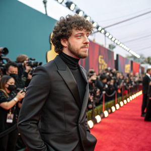 Jack Harlow stands out at a Hollywood award ceremony. His curly hair and beard stand out against the vibrant backdrop. Wearing a sleek black suit, he poses confidently. The setting is filled with flashing cameras and a sea of celebrities.