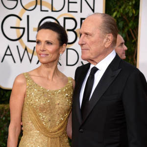 Luciana Pedraza and Robert Duvall at the Golden Globe Awards. Luciana in an elegant gold sequined gown. Robert in a tailored black tuxedo. The backdrop is the iconic Hollywood sign.