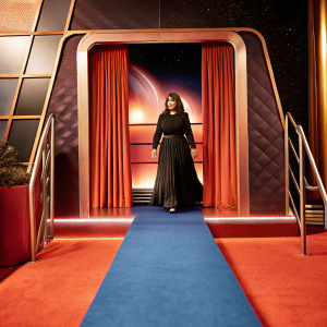Rosie Perez, a Latina actress, dressed in a black long-sleeved dress with a flowing silhouette. Standing confidently on the blue carpeted surface at the 'The Adam Project' premiere. Futuristic themed backdrop with gradient colors and Netflix logo.
