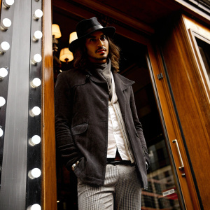 Avan Jogia, sporting long hair and a black hat, at the opening of a John Varvatos store. He is standing in front of the store's facade.