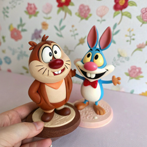 A detailed close-up of a hand holding Ren and Stimpy figurines. The figurines are intricately designed with brown bases and exaggerated comedic expressions. Set against a background of soft pastel floral wallpaper.