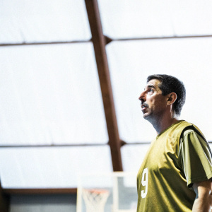 Roy Makaay, in green jersey with number 9, standing still on a dimly lit indoor basketball court.