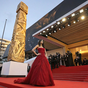 Sofia Boutella at the Cannes Film Festival, wearing a dramatic red gown. She poses in front of a towering, ornate golden statue with intricate carvings. The backdrop is a luxurious red carpet with flashing cameras and intrigued spectators.