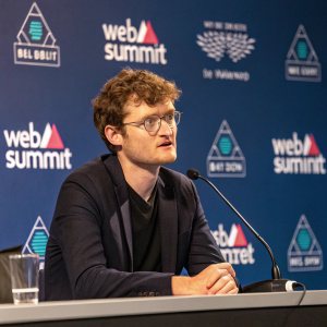 Paddy Cosgrave, CEO and co-founder of Web Summit, addressing the press. Dark suit, seated at a conference table with a microphone in front of him. Web Summit logos and banners in the background.