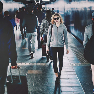 Chloe Grace Moretz walking through a busy metropolitan subway station. Dressed in a casual outfit including a grey hoodie, black yoga pants, and sunglasses. Crowded environment with people and luggage, artificial lighting