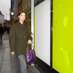 Denise Richards walking down a busy city street at dusk. She's wearing a brown coat and carrying a shiny purple handbag with gold detailing.