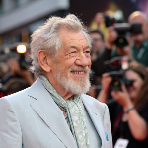 Ian McKellen, with a beard and warm smile, in a light grey suit, at a bustling film festival. Surrounded by flashing cameras and fans.