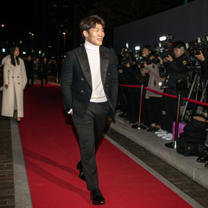 Kim Jong-kook attending a gala event. Elegant attire: black suit and white turtleneck. Red carpet ambiance with flashing cameras and crowd.