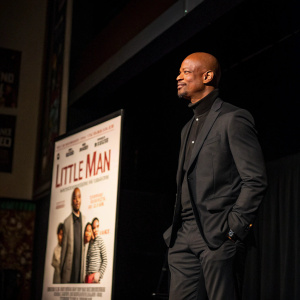 Damon Wayans at the 'Little Man' premiere. Wearing black suit, standing on a dimly lit stage. Movie poster in background.