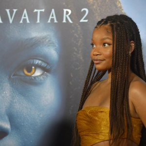 Halle Bailey at the Avatar 2 premiere. Wearing a golden top, long braided hair, standing confidently in front of a blue backdrop with text elements.