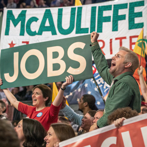 Crowd at political rally, enthusiastic supporters holding banners. McAuliffe and jobs in large letters on signs.
