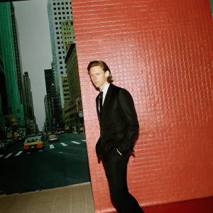 Tom Hiddleston, dressed in a sleek black suit and tie, standing in front of a bustling cityscape.
