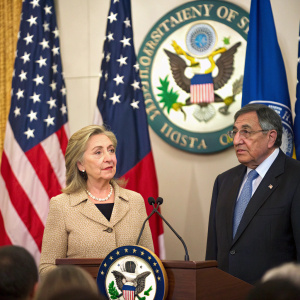 Hillary Clinton and Leon Panetta addressing a crowded press conference. Hillary Clinton, wearing a beige blazer, stands alongside Leon Panetta in a dark suit. Behind them, the Department of State emblem is prominently displayed. American and other national flags flutter in the background.