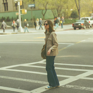 Anne Hathaway on a bustling city street, wearing casual attire. Dark blue jeans, light brown jacket, casual sneakers. No backdrop logos