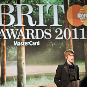 Justin Bieber at the BRIT Awards 2011. Caucasian male, young, casual hairstyle, in a black tuxedo. Large backdrop with 'BRIT AWARDS 2011 with MasterCard' text. Live event moment.