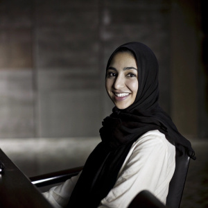 Photorealistic image of a young professional Asian woman, fair skin, mid-20s, wearing a black hijab and white blouse. Warm smile, in a sleek modern workspace. Background blurred, focus on upper body.