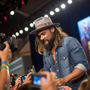 Jason Momoa at a comic-con, interacting with fans. Wearing a gray fedora, denim shirt. Bright lights and camera flashes