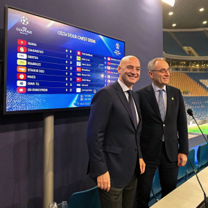 Gianni Infantino and Giorgio Marchetti, in suits, during a UEFA Champions League press conference. Large screen displays tournament standings.