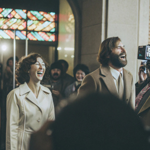 Kristen Schaal and Will Forte laughing together at a bustling after party, surrounded by fans and media. Neon lights in the background.