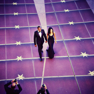 Terrence Howard and Mira Pak walking on a star-studded red carpet. Wearing black gowns and tuxedos. Paparazzi snapping photos.