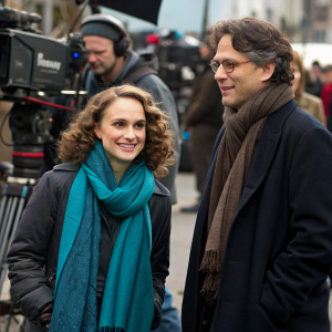 Natalie Portman, with curly brown hair and wearing a teal scarf and jacket, alongside Scott Cohen in a dark brown scarf and dark jacket. Both are on a bustling outdoor film set.