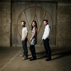 Aaron Paul, Lisa Joy, and Min Jin Lee at a Westworld promotional event. They are dressed in casual attire, with Aaron Paul wearing a light-colored shirt, Lisa Joy in a dark ensemble, and Min Jin Lee in white. Aaron Paul is bowing slightly, while they stand against a backdrop featuring large promotional logos.