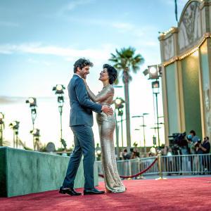 Matt Dallas and Jaimie Alexander at a Hollywood premiere. Matt in a navy blue suit, Jaimie in a shimmering gold gown. Flashing cameras and red carpet in the background.